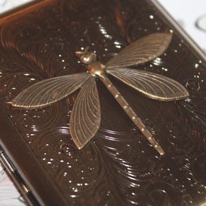 May include: A bronze cigarette case with an embossed floral pattern and a decorative dragonfly. The dragonfly is made of bronze-coloured metal and is centered on the case. The case has a rectangular shape.