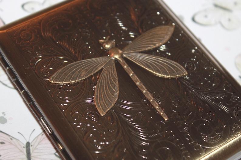 May include: A bronze cigarette case with an embossed floral pattern and a decorative dragonfly. The dragonfly is made of bronze-coloured metal and is centered on the case. The case has a rectangular shape.