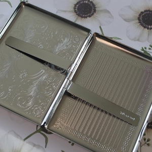 May include: A silver cigarette case with an engraved floral design on one side and a textured surface on the other. The case has a metal hinge and a slide-out mechanism for holding cigarettes.