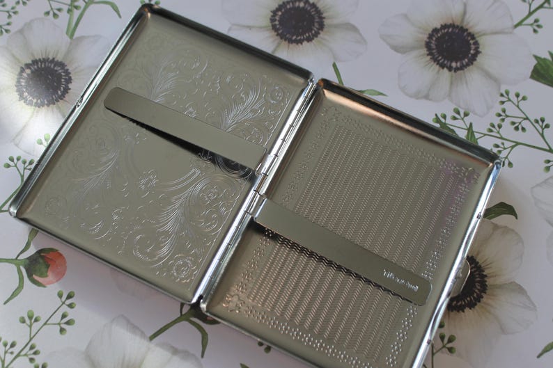 May include: A silver cigarette case with an engraved floral design on one side and a textured surface on the other. The case has a metal hinge and a slide-out mechanism for holding cigarettes.