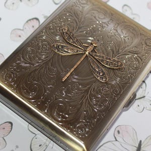 Puede incluir: Una pitillera rectangular de metal con acabado en lat&oacute;n antiguo, que presenta un intrincado patr&oacute;n floral en relieve en toda la superficie y un destacado emblema de lib&eacute;lula tridimensional centrado en la tapa frontal.
