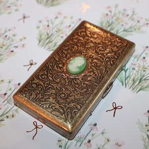 May include: A gold metal cigarette case with an ornate floral design and a green cameo in the center.