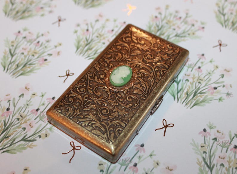 May include: A gold metal cigarette case with an ornate floral design and a green cameo in the center.