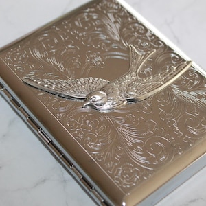 Puede incluir: Cajetilla de cigarrillos de metal plateado con un dise&ntilde;o de p&aacute;jaro grabado. La cajetilla presenta un detallado patr&oacute;n floral y un p&aacute;jaro en relieve en el centro. La cajetilla es rectangular y tiene una tapa con bisagras.