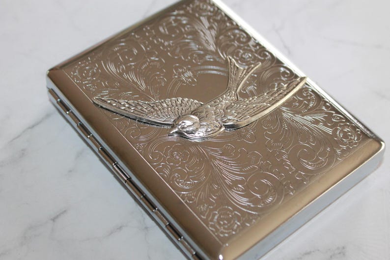 Puede incluir: Cajetilla de cigarrillos de metal plateado con un dise&ntilde;o de p&aacute;jaro grabado. La cajetilla presenta un detallado patr&oacute;n floral y un p&aacute;jaro en relieve en el centro. La cajetilla es rectangular y tiene una tapa con bisagras.