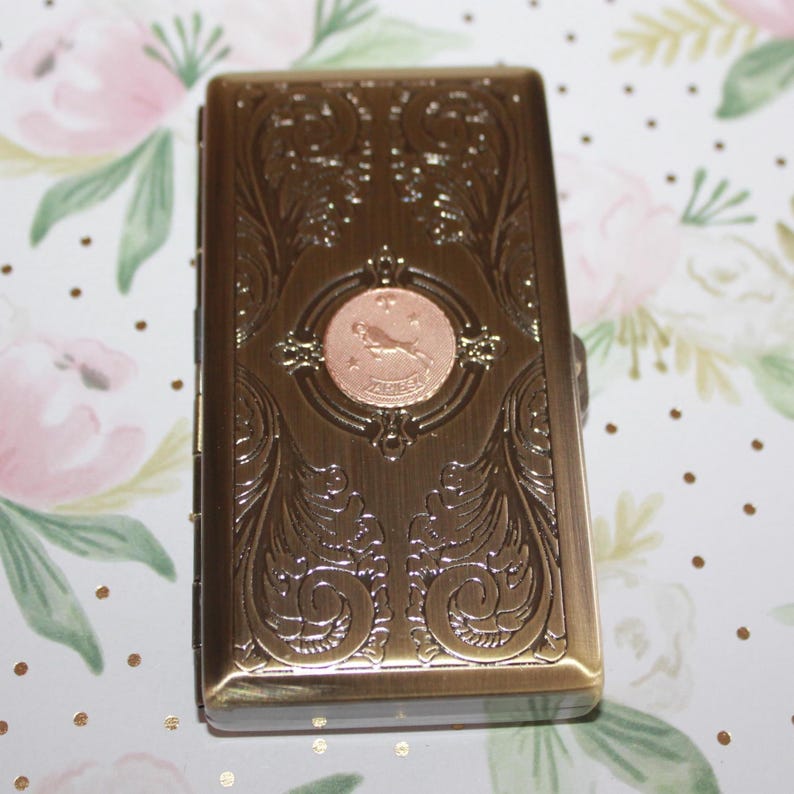 May include: A rectangular, antique-style metal case with an ornate, embossed floral design. The case is bronze-colored with a copper-colored circular emblem featuring a bird. The case is closed and has a hinged lid.