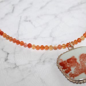 May include: A necklace featuring a strand of small, round, translucent orange beads. A square-shaped pendant with rounded corners hangs from the strand, displaying a detailed red dragon illustration on a white background, set in a silver-toned metal frame.