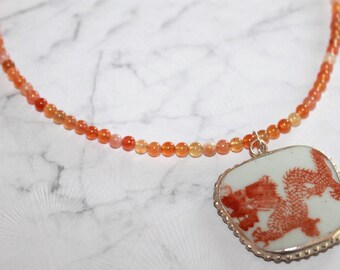 May include: A necklace featuring a strand of small, round, translucent orange beads. A square-shaped pendant with rounded corners hangs from the strand, displaying a detailed red dragon illustration on a white background, set in a silver-toned metal frame.