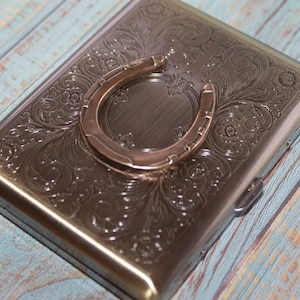 Puede incluir: Un estuche metálico rectangular con un patrón floral en relieve y un acento de herradura. El estuche tiene una tapa con bisagras y un acabado plateado. La herradura es de un tono plateado más claro.