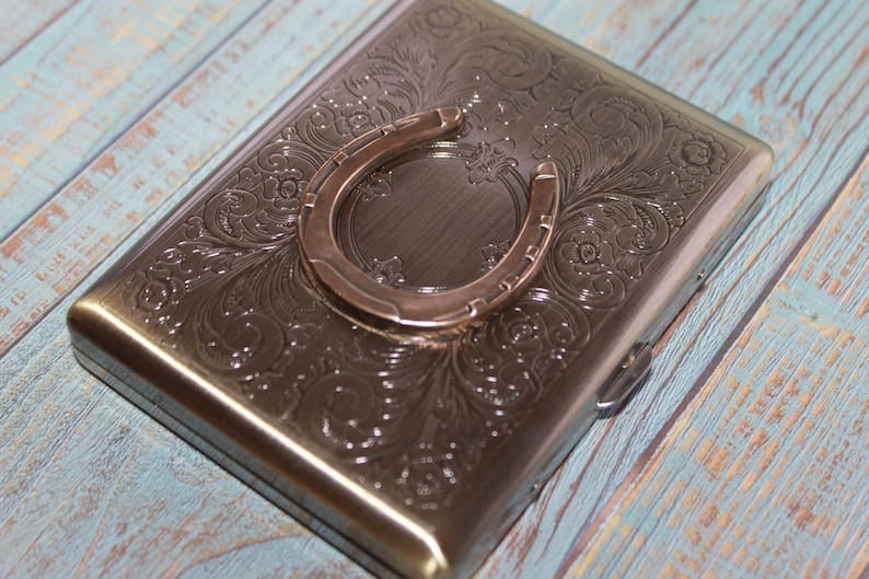 May include: A rectangular, metallic case with an embossed floral pattern and a horseshoe accent. The case has a hinged lid and a silver finish. The horseshoe is a lighter shade of silver.