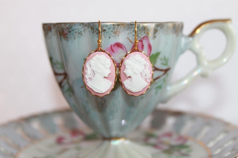 May include: A pair of gold earrings with pink and white cameo portraits. The cameos are oval shaped and feature a woman's profile.