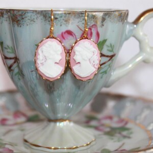 May include: A pair of gold earrings with pink cameo portraits. The earrings are hanging from a vintage teacup.