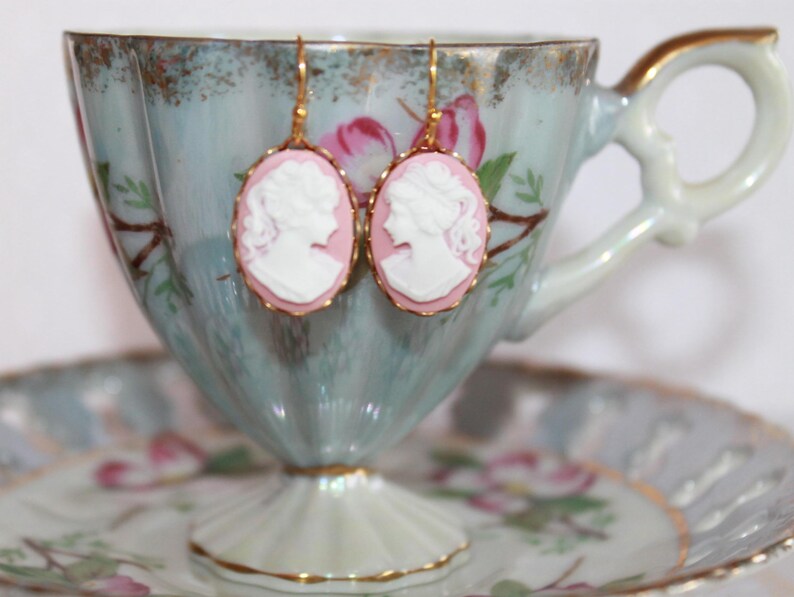 May include: A pair of gold earrings with pink cameo portraits. The earrings are hanging from a vintage teacup.