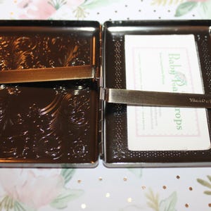 May include: Open, dark brown metal case with ornate floral detailing. The case is open, revealing a card with the text "Baby Tea Drops". The case has a bronze-colored clasp.