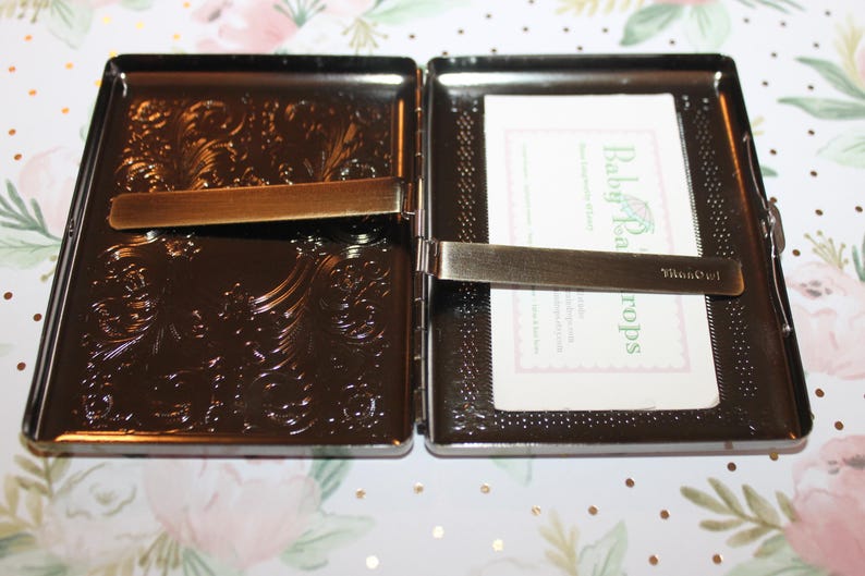 May include: Open, dark brown metal case with ornate floral detailing. The case is open, revealing a card with the text "Baby Tea Drops". The case has a bronze-colored clasp.