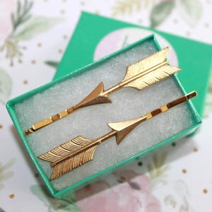 May include: Two gold-toned metal hair pins shaped like arrows, featuring textured fletching and pointed tips. The hair pins are resting inside a small, rectangular, bright green cardboard box lined with white foam padding for display.