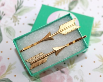 May include: Two gold-toned metal hair pins shaped like arrows, featuring textured fletching and pointed tips. The hair pins are resting inside a small, rectangular, bright green cardboard box lined with white foam padding for display.