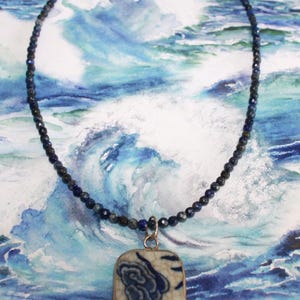 May include: A necklace featuring a strand of small, faceted dark blue beads and a rectangular pendant. The pendant has a cream-colored base with a blue floral pattern and a visible crackle glaze finish, suspended from a silver-toned metal bail.