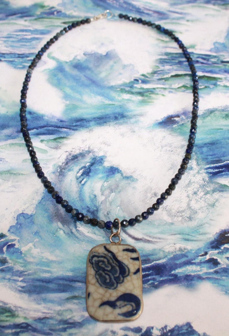 May include: A necklace featuring a strand of small, faceted dark blue beads and a rectangular pendant. The pendant has a cream-colored base with a blue floral pattern and a visible crackle glaze finish, suspended from a silver-toned metal bail.