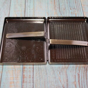 May include: An open, rectangular, dark brown metal cigarette case with a decorative floral pattern on one side and a striped pattern on the other. Two metal clips are visible. The case is on a light blue and brown wooden surface.