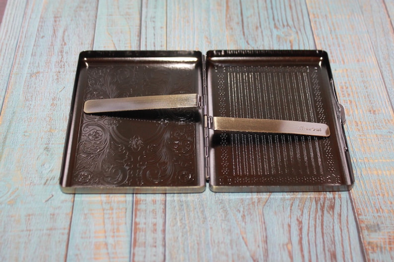 May include: An open, rectangular, dark brown metal cigarette case with a decorative floral pattern on one side and a striped pattern on the other. Two metal clips are visible. The case is on a light blue and brown wooden surface.