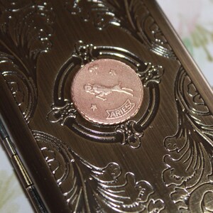 May include: A bronze-colored metal case with an embossed floral design. A circular copper-colored emblem in the center features the Aries zodiac symbol and the word "ARIES". The case has a hinged closure.