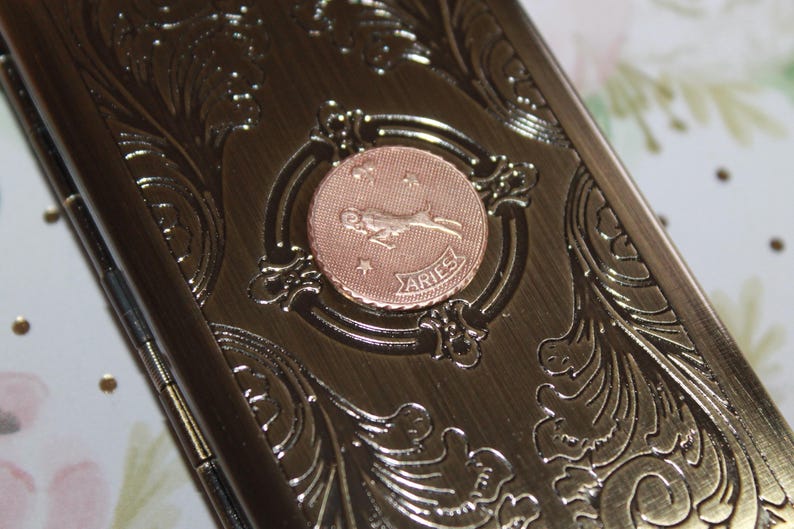 May include: A bronze-colored metal case with an embossed floral design. A circular copper-colored emblem in the center features the Aries zodiac symbol and the word "ARIES". The case has a hinged closure.
