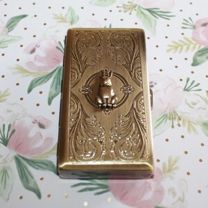 Puede incluir: Un estuche rectangular de metal en tono lat&oacute;n antiguo que presenta un patr&oacute;n de volutas intrincado y en relieve en su superficie. Centrada en la parte frontal hay una peque&ntilde;a figura tridimensional de un conejo sentado dentro de un marco circular.