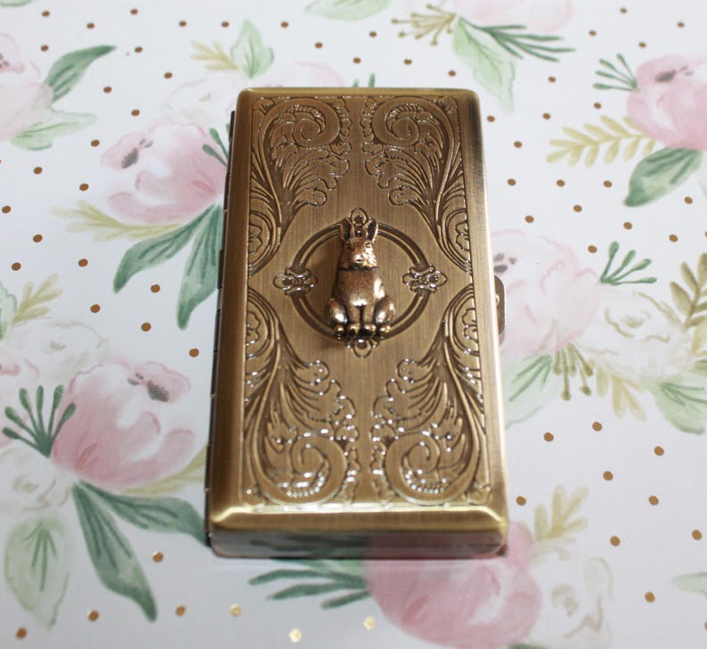 Puede incluir: Un estuche rectangular de metal en tono lat&oacute;n antiguo que presenta un patr&oacute;n de volutas intrincado y en relieve en su superficie. Centrada en la parte frontal hay una peque&ntilde;a figura tridimensional de un conejo sentado dentro de un marco circular.
