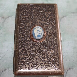 Puede incluir: Un estuche rectangular de estilo antiguo con un detallado patr&oacute;n floral en tonos bronce y plata. Un peque&ntilde;o camafeo ovalado con fondo azul e imagen blanca est&aacute; centrado en la parte delantera. El camafeo est&aacute; enmarcado en un metal color cobre. El estuche se utiliza probablemente para guardar objetos peque&ntilde;os.