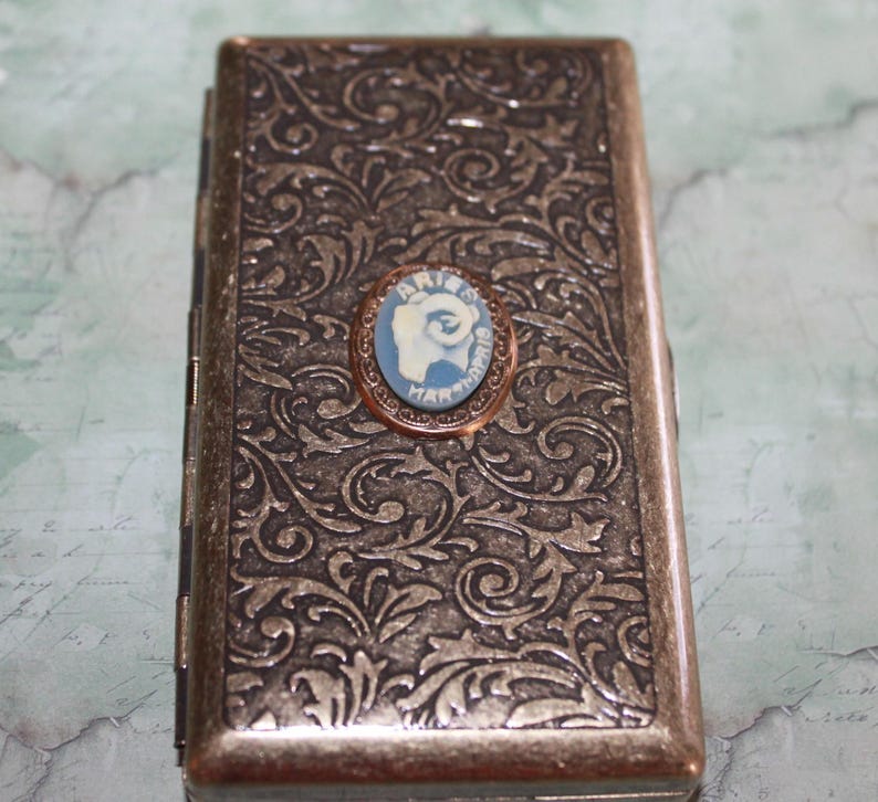 Puede incluir: Un estuche rectangular de estilo antiguo con un detallado patr&oacute;n floral en tonos bronce y plata. Un peque&ntilde;o camafeo ovalado con fondo azul e imagen blanca est&aacute; centrado en la parte delantera. El camafeo est&aacute; enmarcado en un metal color cobre. El estuche se utiliza probablemente para guardar objetos peque&ntilde;os.