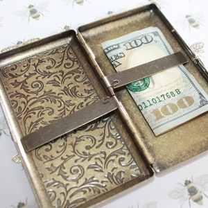 May include: A gold-toned metal cigarette case with an ornate floral design. The case is open and a $100 bill is inside.