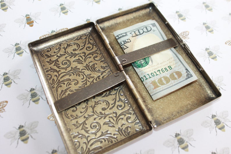 May include: A gold-toned metal cigarette case with an ornate floral design. The case is open and a $100 bill is inside.