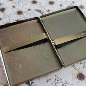 May include: A rectangular metal cigarette case with a polished silver finish, opened to reveal two interior compartments. Each side features an engraved floral pattern and a horizontal spring-loaded metal bar for securing contents. The word "Mascot" is etched on one bar.