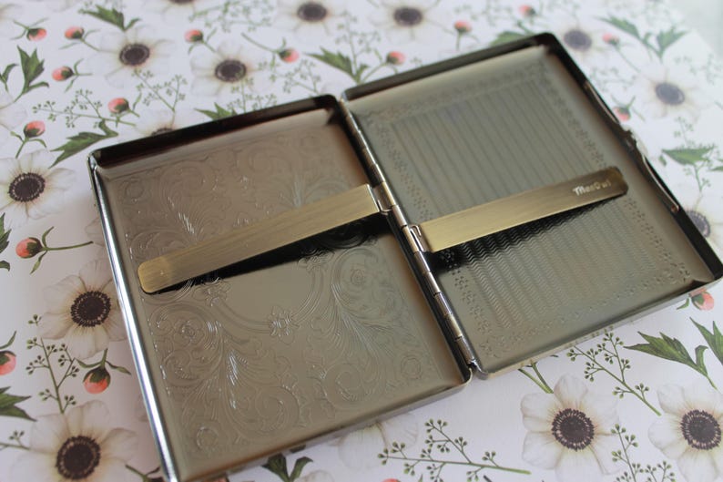May include: A rectangular metal cigarette case with a polished silver finish, opened to reveal two interior compartments. Each side features an engraved floral pattern and a horizontal spring-loaded metal bar for securing contents. The word "Mascot" is etched on one bar.