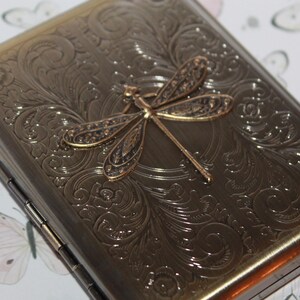 Puede incluir: Una pitillera rectangular de metal en tono bronce con un intrincado patr&oacute;n floral en relieve en toda su superficie. Un prominente emblema tridimensional de lib&eacute;lula con alas texturizadas se encuentra centrado en la tapa frontal.