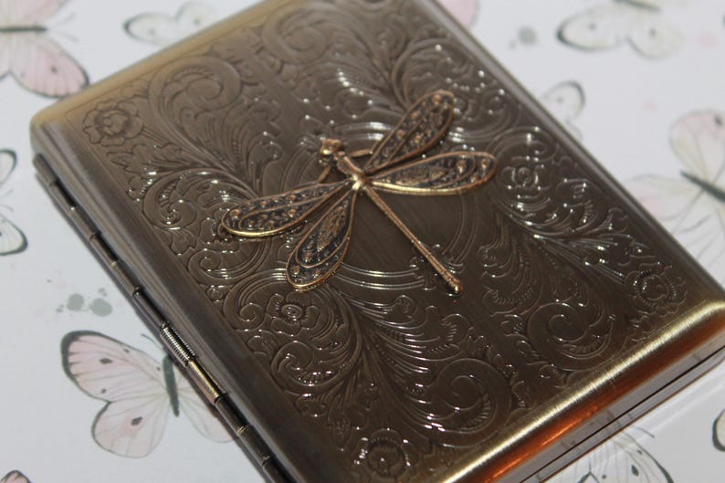 Puede incluir: Una pitillera rectangular de metal en tono bronce con un intrincado patr&oacute;n floral en relieve en toda su superficie. Un prominente emblema tridimensional de lib&eacute;lula con alas texturizadas se encuentra centrado en la tapa frontal.