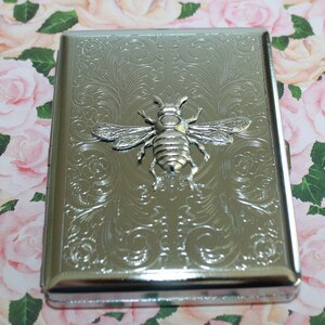 May include: Silver metal cigarette case with an embossed bee design. The case has a rectangular shape with rounded edges and a decorative floral pattern. The bee is in the centre of the case. The background is a floral pattern.
