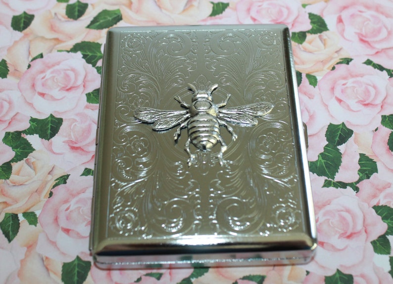 May include: Silver metal cigarette case with an embossed bee design. The case has a rectangular shape with rounded edges and a decorative floral pattern. The bee is in the centre of the case. The background is a floral pattern.