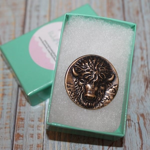 May include: A round, bronze-colored metal pin featuring a detailed depiction of a bison's head. The pin is presented inside a small, open, mint-green gift box with white padding. The box sits on a light-colored, weathered wooden surface.