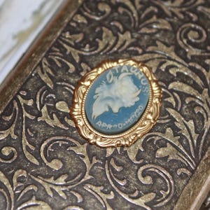 May include: An antique gold-toned metal case with an ornate floral design. A gold-framed cameo with a blue background and a white profile is centered on the case. The cameo reads "TAURUS APR 20-MAY 20".