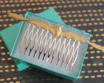 Op de afbeelding: Een goudkleurige haarkam met een gedetailleerd vleermuisontwerp op de bovenste balk. De kam heeft negen slanke, gebogen tanden die vanaf de vleugels van de vleermuis naar beneden lopen. Het accessoire wordt gepresenteerd in een kleine, turquoise geschenkdoos.