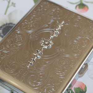 May include: A silver cigarette case with an ornate floral design and a silver flower accent.