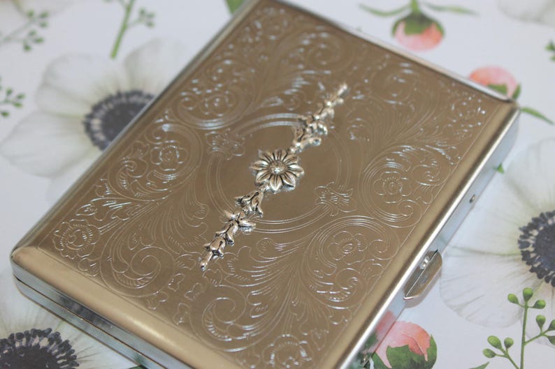 May include: A silver cigarette case with an ornate floral design and a silver flower accent.