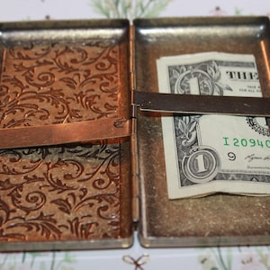 May include: A silver metal cigarette case with a floral pattern on the front. The case is open and a dollar bill is visible inside.