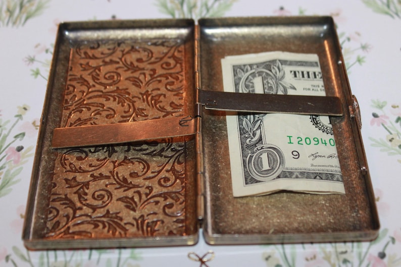 May include: A silver metal cigarette case with a floral pattern on the front. The case is open and a dollar bill is visible inside.