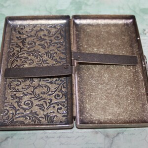 Puede incluir: Estuche de cigarrillos de metal antiguo abierto. El lado izquierdo presenta un dise&ntilde;o floral en relieve ornamentado. El lado derecho tiene una apariencia de metal cepillado texturizado. El estuche est&aacute; abierto, revelando el interior. Art&iacute;culo vintage.