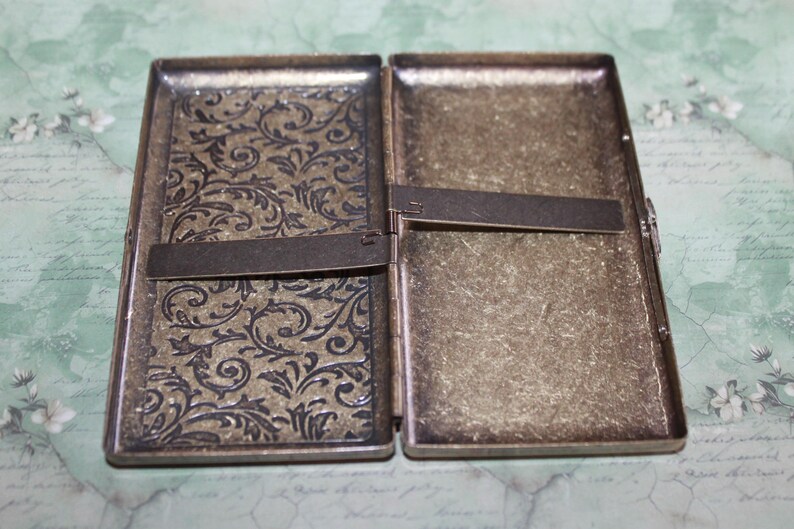 Puede incluir: Estuche de cigarrillos de metal antiguo abierto. El lado izquierdo presenta un dise&ntilde;o floral en relieve ornamentado. El lado derecho tiene una apariencia de metal cepillado texturizado. El estuche est&aacute; abierto, revelando el interior. Art&iacute;culo vintage.