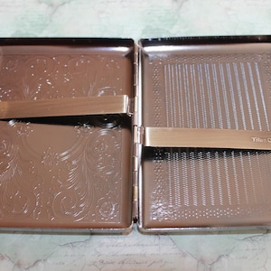 Puede incluir: Una pitillera rectangular de metal en posici&oacute;n abierta con un acabado de bronce pulido. El interior cuenta con dos barras met&aacute;licas con resorte para asegurar el contenido. El lado izquierdo tiene un patr&oacute;n floral en relieve, mientras que el lado derecho presenta un patr&oacute;n de chevr&oacute;n texturizado y el texto grabado TrimOwl.
