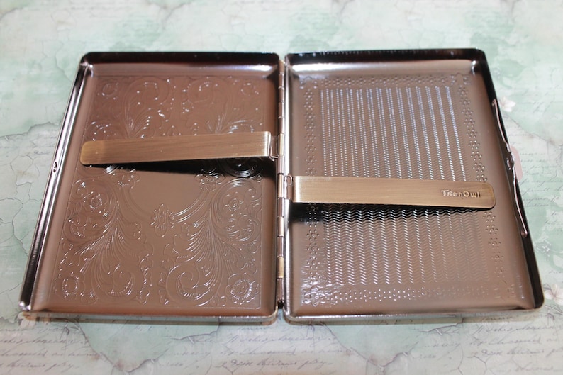 Puede incluir: Una pitillera rectangular de metal en posici&oacute;n abierta con un acabado de bronce pulido. El interior cuenta con dos barras met&aacute;licas con resorte para asegurar el contenido. El lado izquierdo tiene un patr&oacute;n floral en relieve, mientras que el lado derecho presenta un patr&oacute;n de chevr&oacute;n texturizado y el texto grabado TrimOwl.